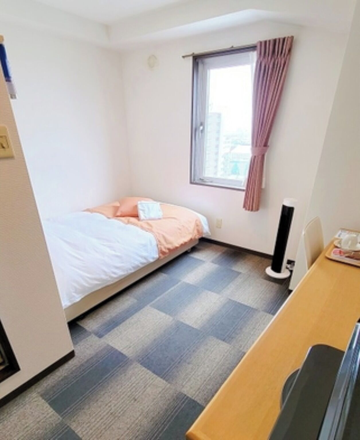 Ein kleines Hotelzimmer mit einem Einzelbett, einem Schreibtisch, einem Stuhl, einem Fenster mit Vorhängen und einer Heizung.