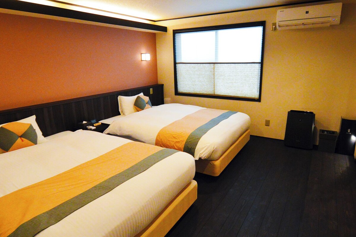 A hotel room with two beds, a window with blinds, an air conditioner, and a desk with a speaker.
