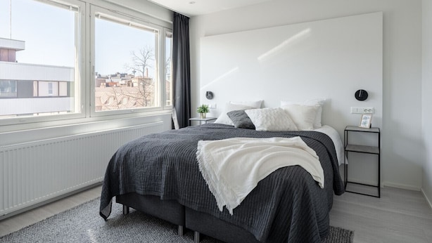 Een moderne slaapkamer met een groot bed, een nachtkastje, een raam en uitzicht op gebouwen.