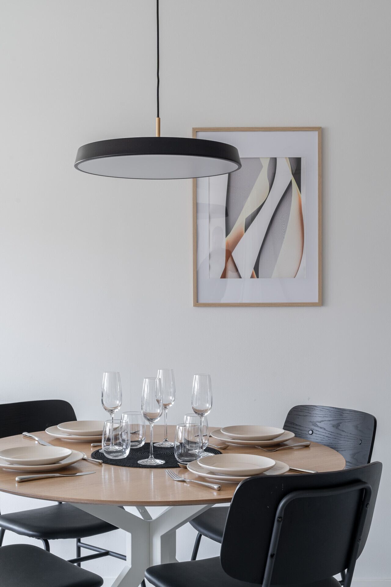A dining area with a round table set for six, black chairs, a pendant light, and a framed abstract painting on the wall.