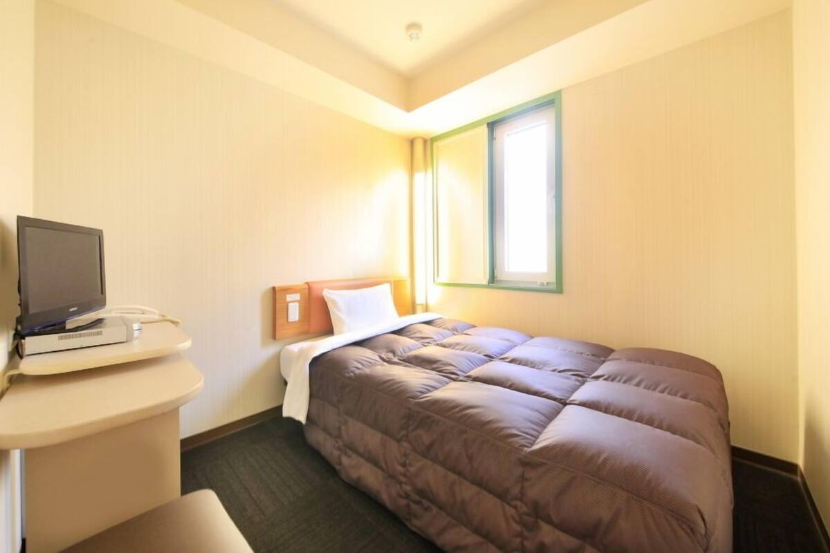 A hotel room with a bed, a desk with a computer, a window, and a door.