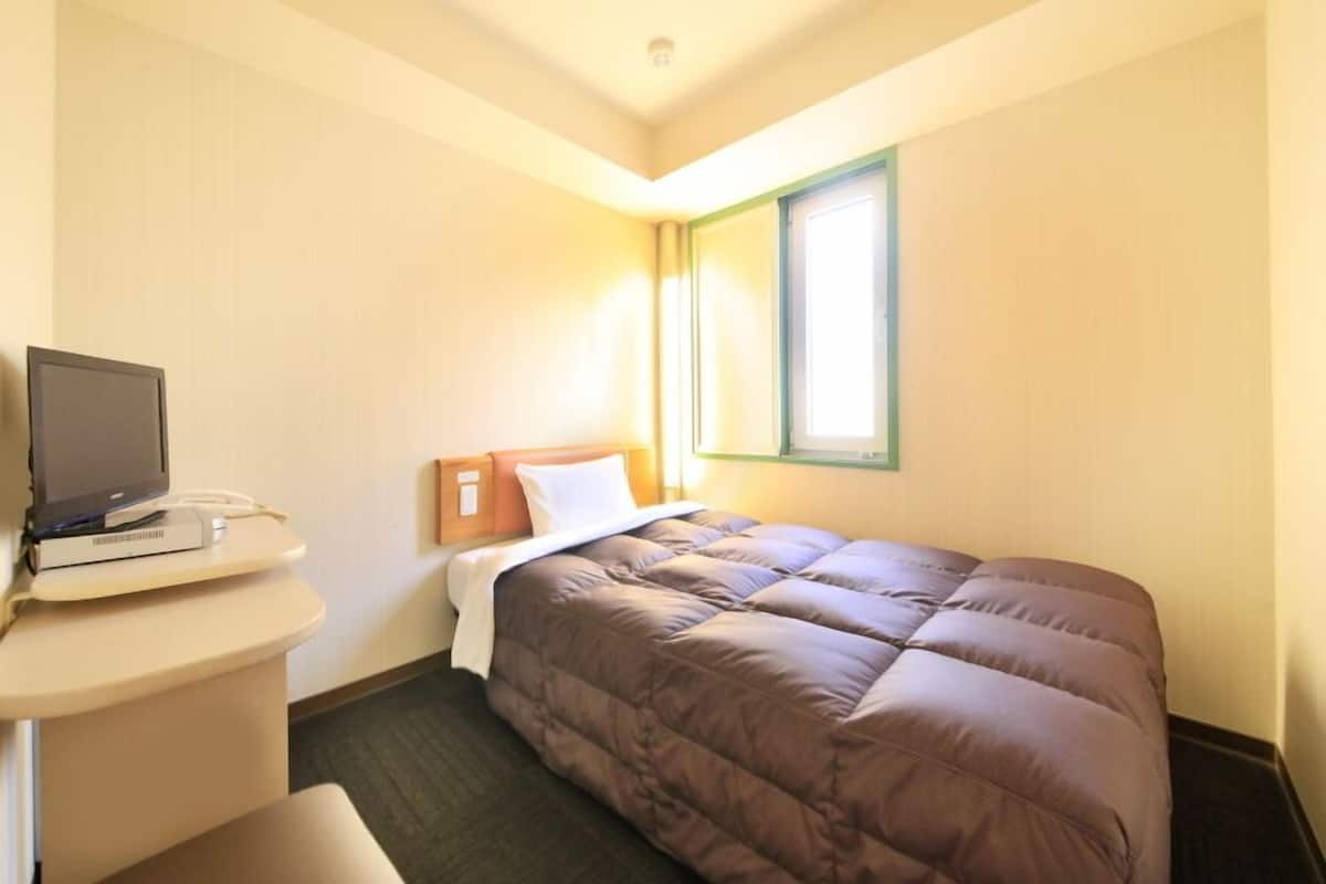 A hotel room with a bed, a desk with a computer, a window, and a door.