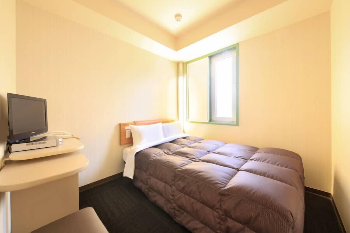 A hotel room with a bed, a desk with a television and phone, and a window.
