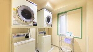Uma lavanderia com duas máquinas de lavar, uma secadora e um carrinho com produtos de lavagem.