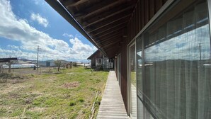 A covered outdoor area with a wooden deck, a glass door, and a view of a grassy field and distant buildings. - Tabinoyado Tenohirani Taiyonoie (Tome)