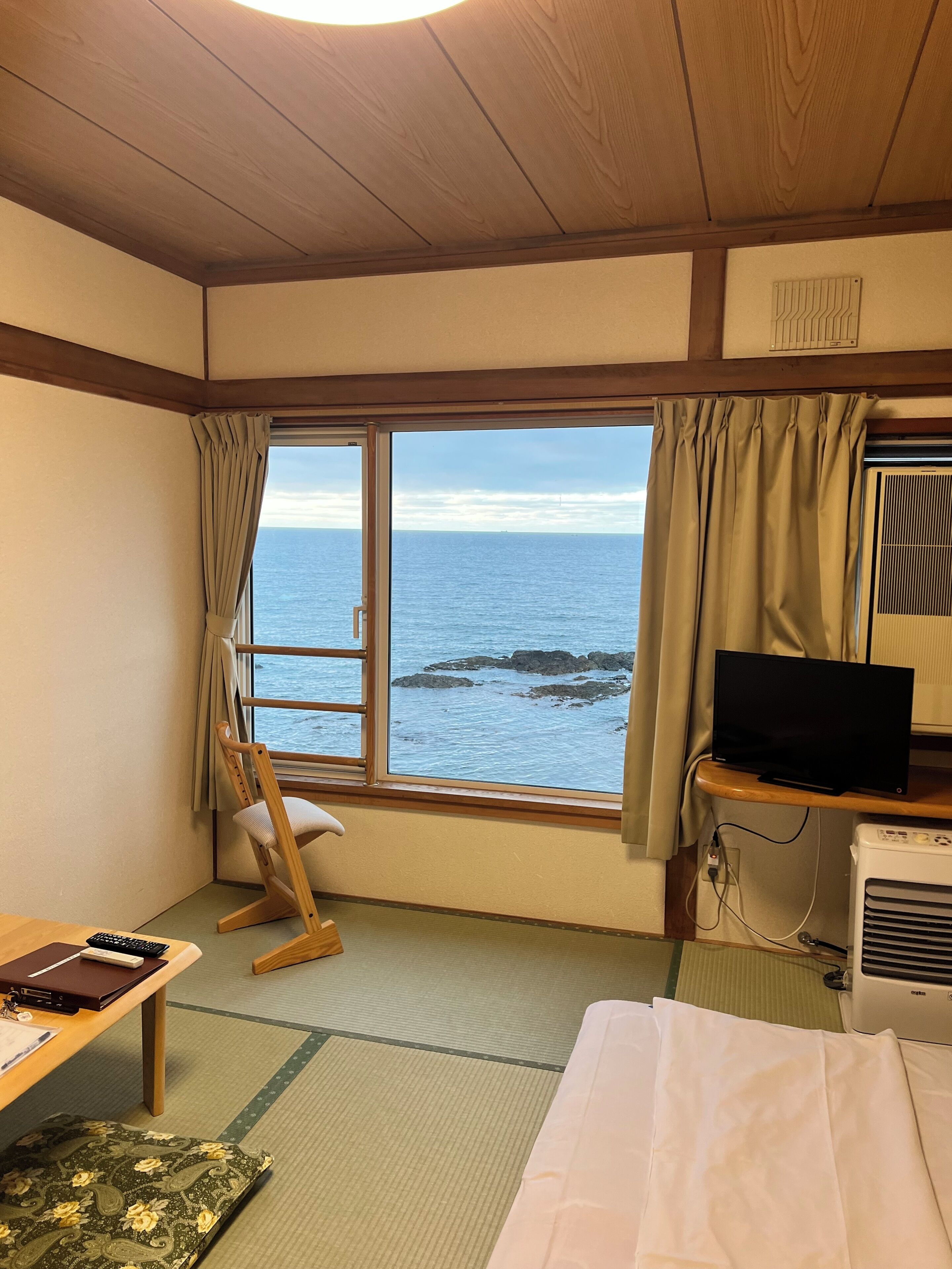 A room with a large window, a television, a desk with a laptop, and a bed.