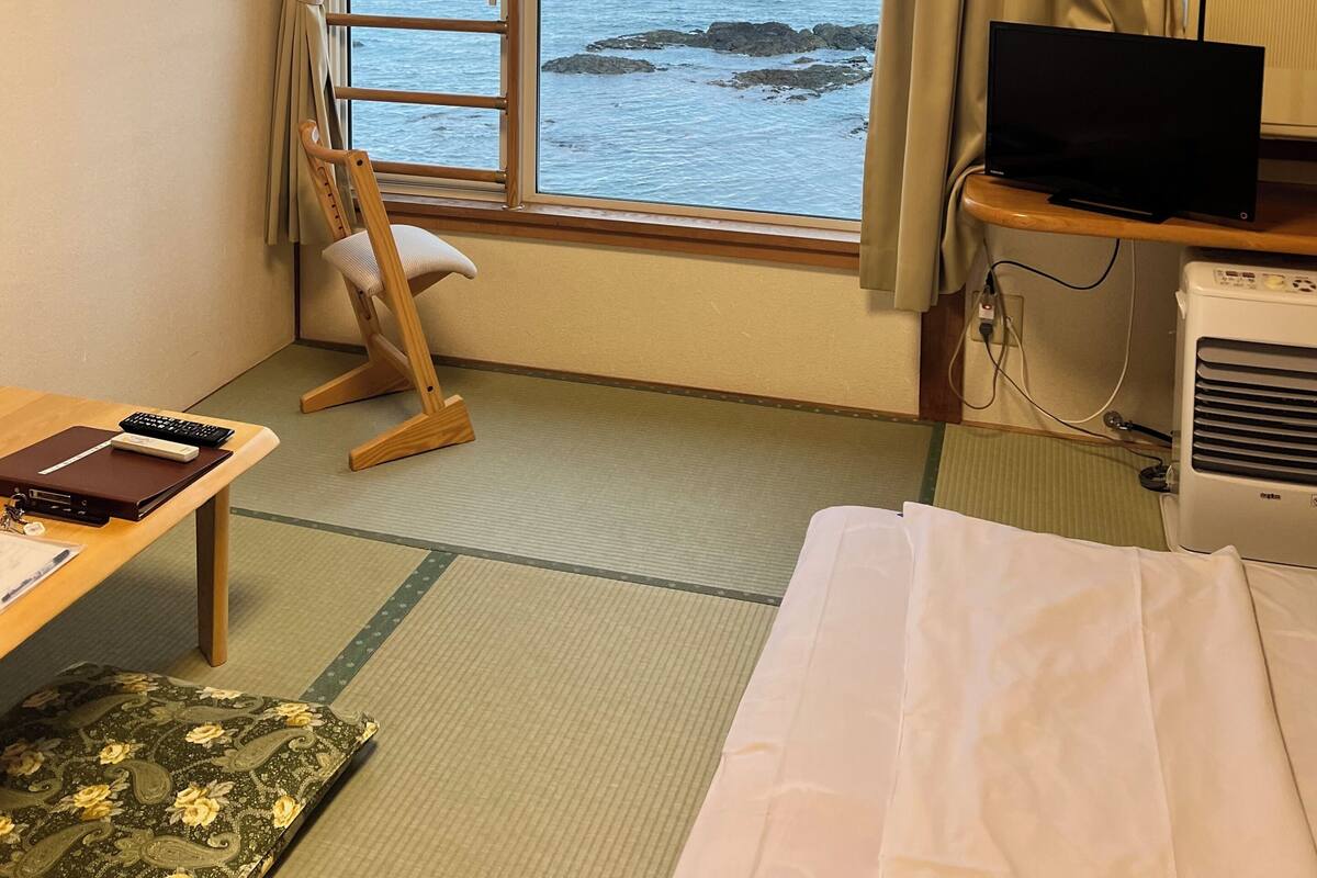 Een kamer met een groot raam, een televisie, een bureau met een laptop en een bed.