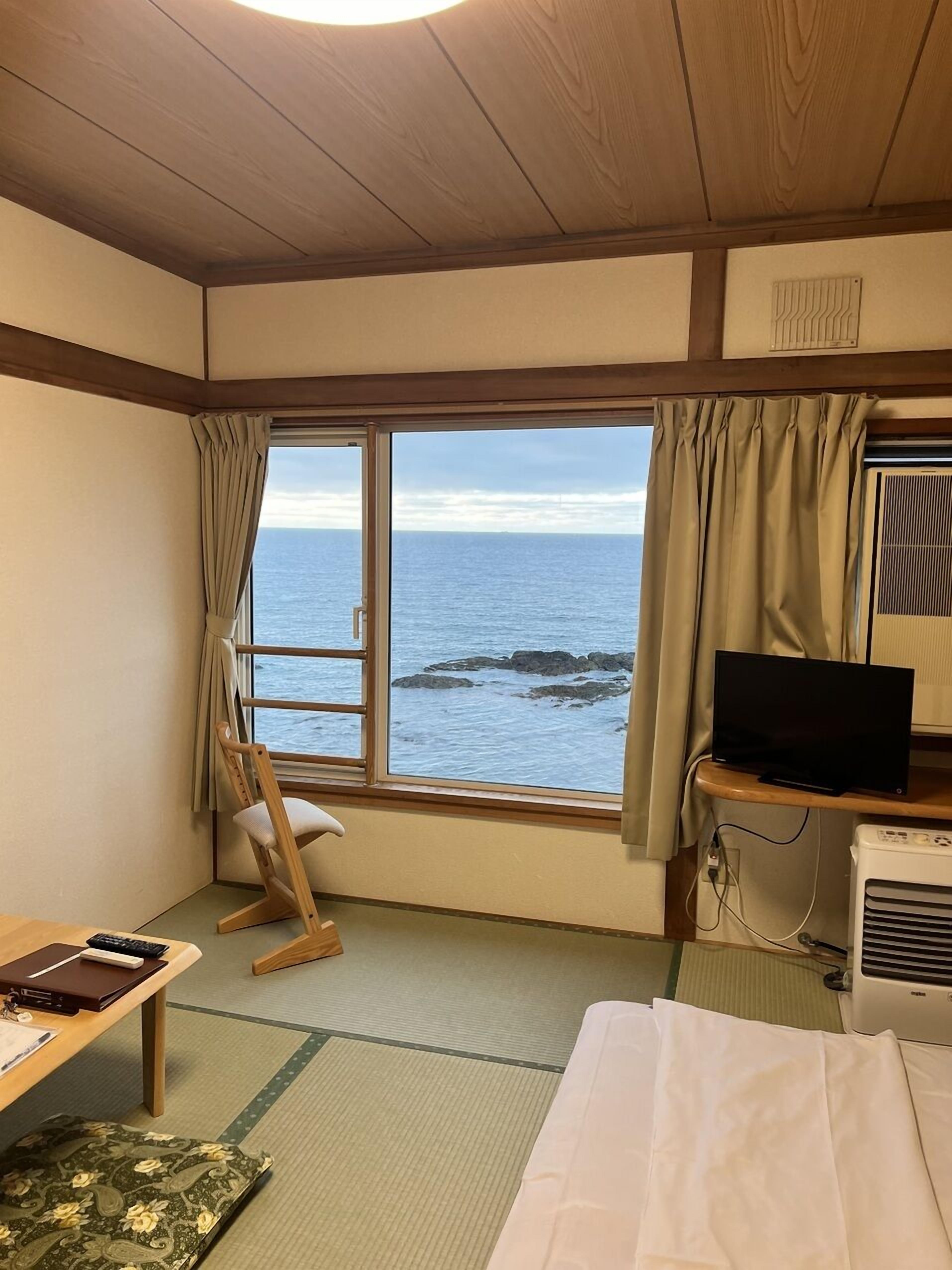 A room with a bed, a desk, a chair, a TV, and a window with a view of the ocean.