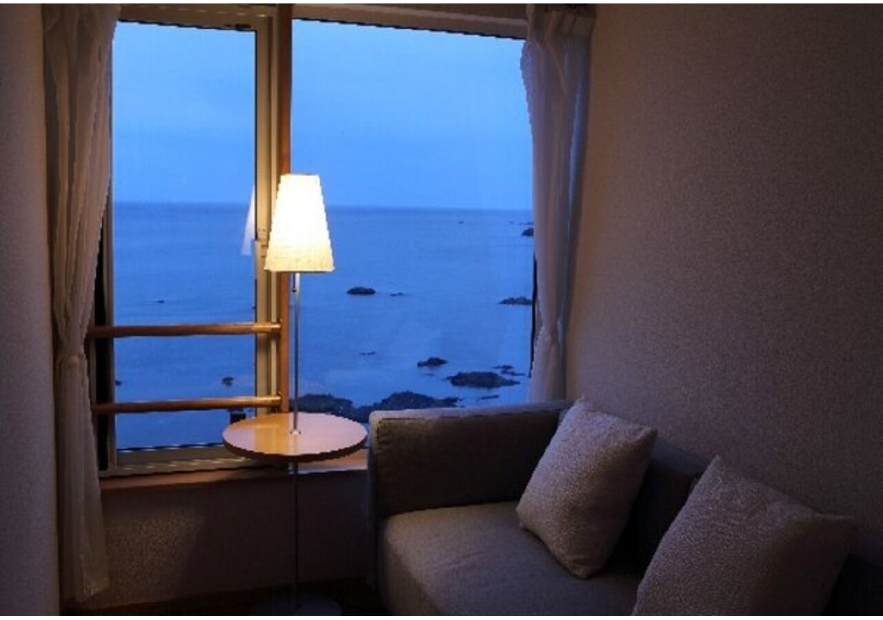 A room with a view of the sea through a window, a lamp on a small table, and a sofa with cushions.