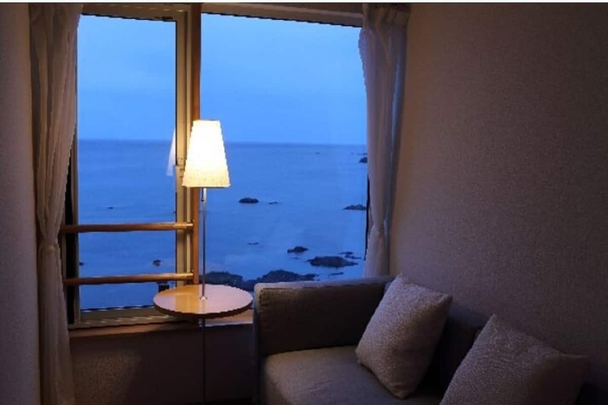 Een kamer met uitzicht op zee door een raam, een lamp op een klein tafeltje en een bank met kussens.