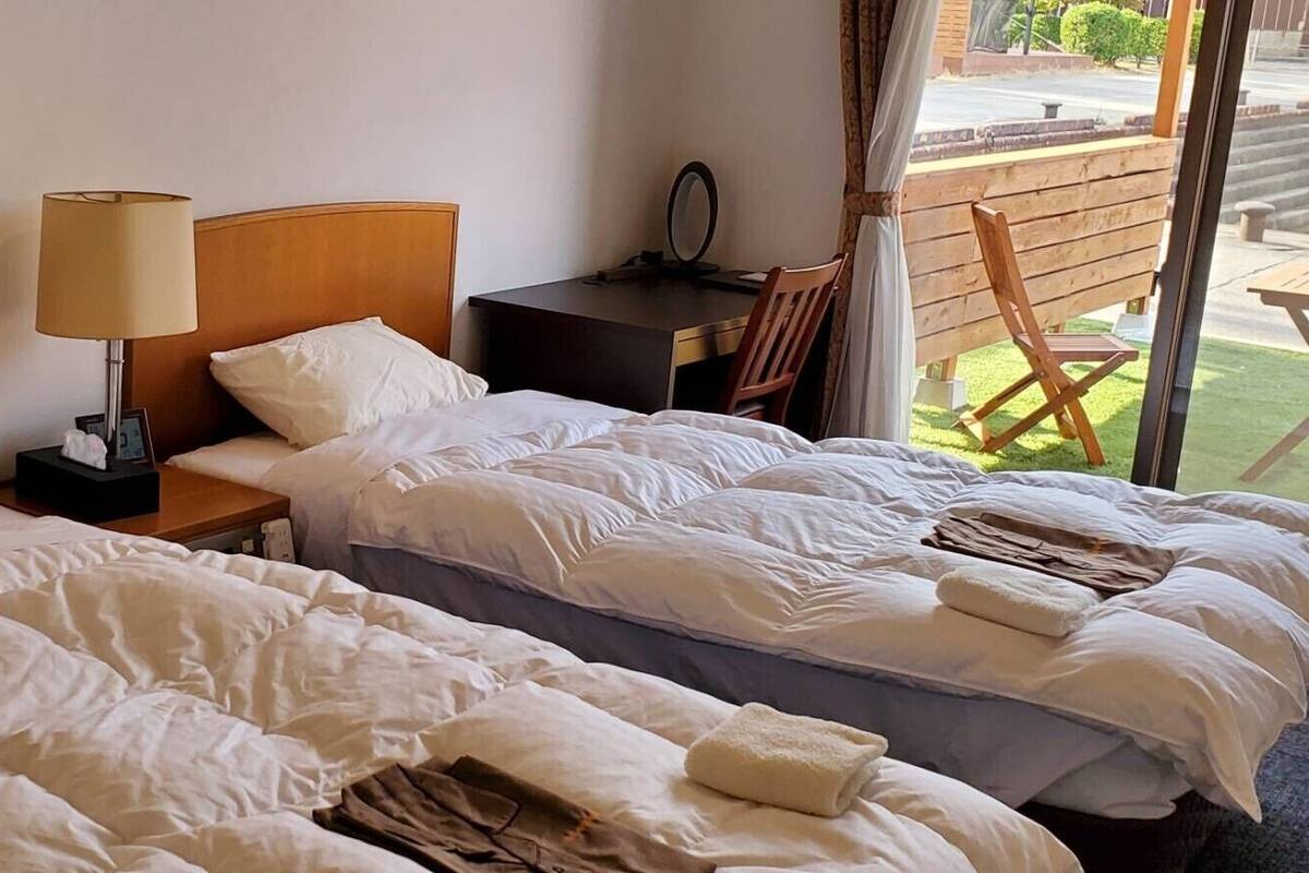 Quarto de hotel com duas camas, uma mesa de cabeceira com luminária, uma pequena escrivaninha e uma cadeira.