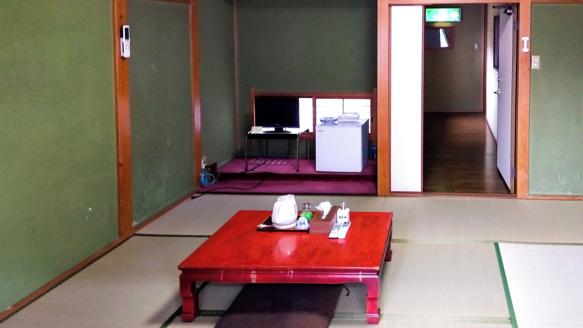 Ett traditionellt japanskt rum med tatamimattor på golvet, ett rött bord med ett teservis, en tv och en skjutdörr som leder till ett annat rum.