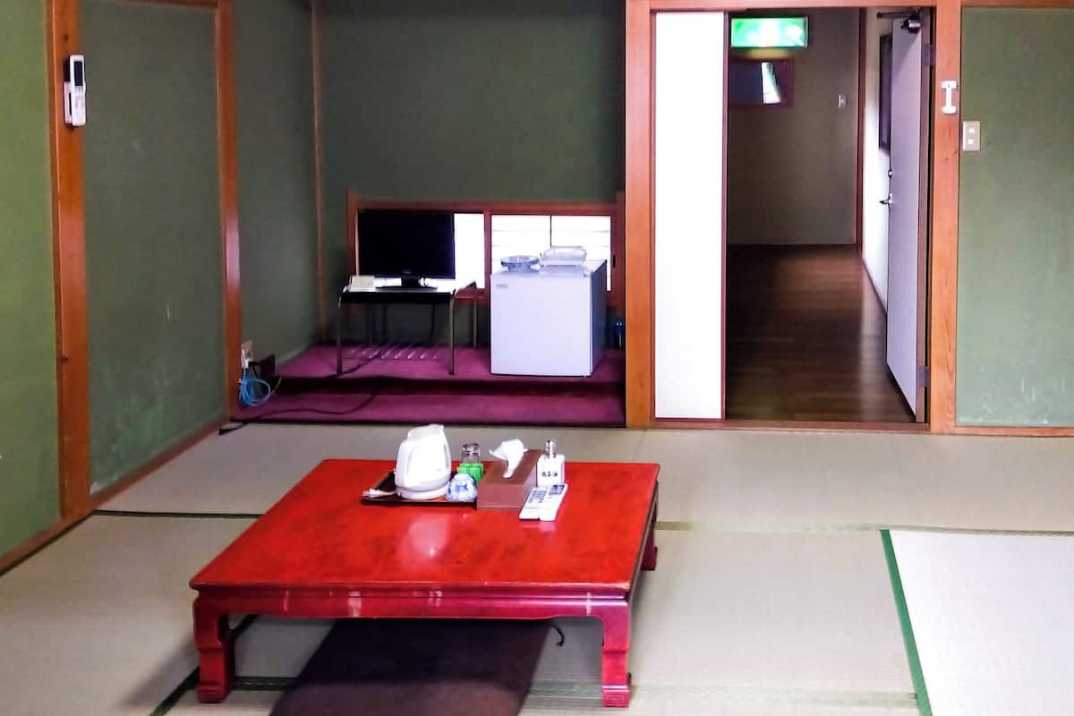A traditional Japanese room with tatami flooring, a red table with a tea set, a television, and a sliding door leading to another room.