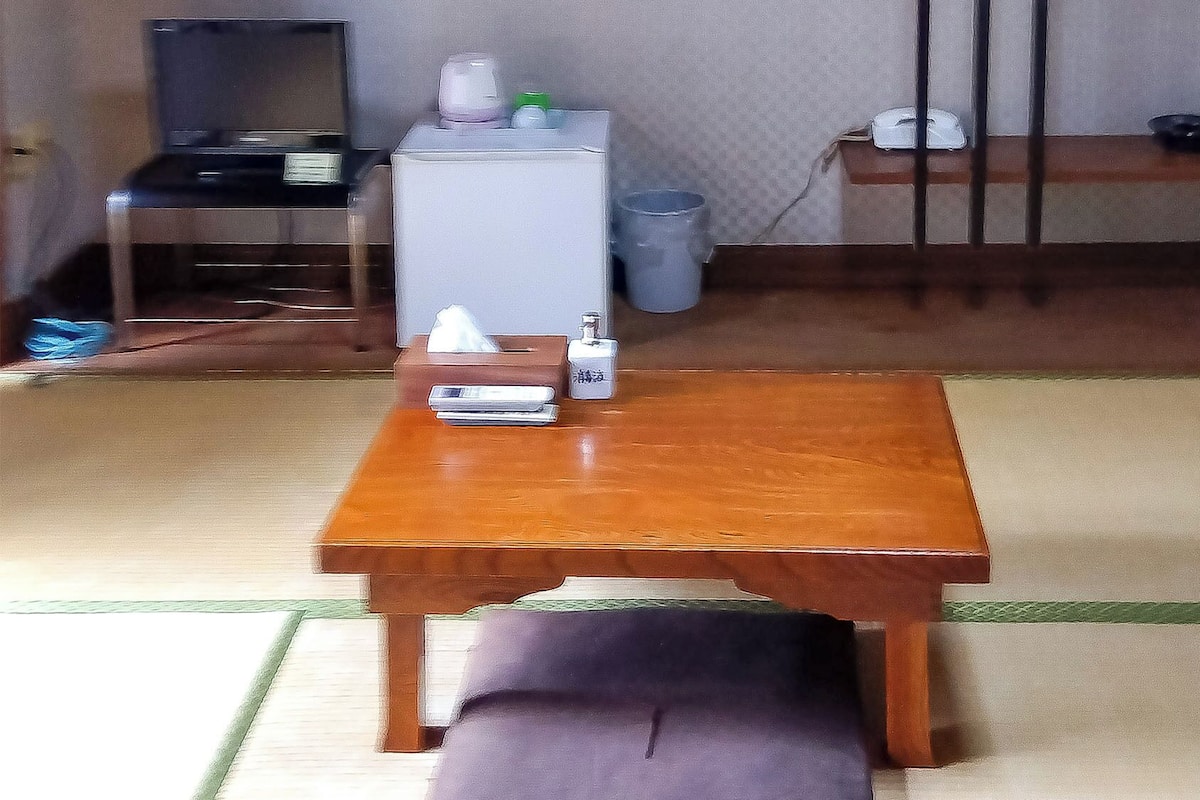 A room with a wooden table, a trash bin, a small shelf, and a television.