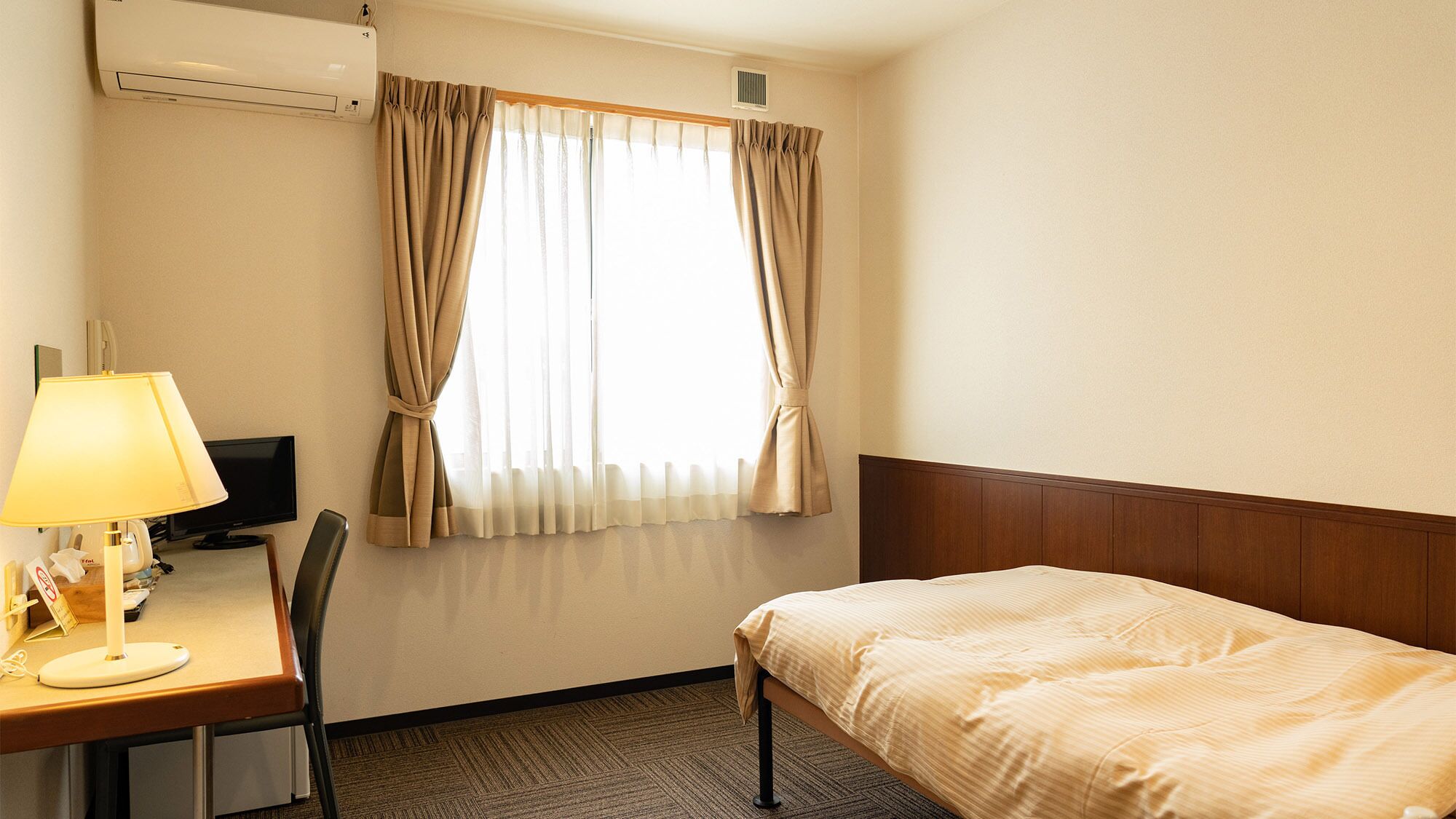 Une chambre d’hôtel avec un lit, un bureau avec une lampe, une télévision et une fenêtre avec des rideaux.