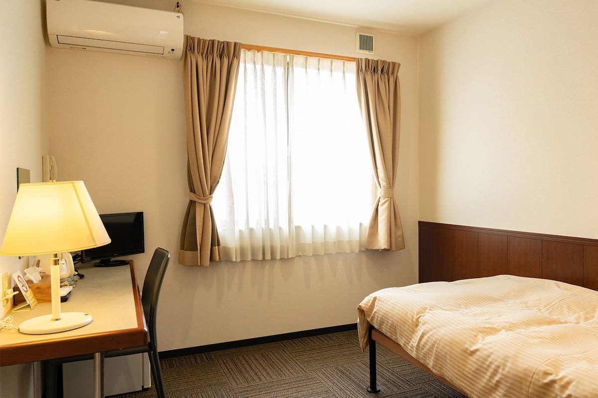 A hotel room with a bed, a desk with a lamp, a television, and a window with curtains.