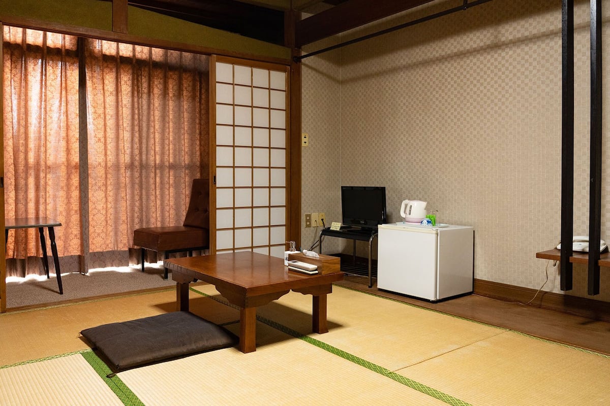 A traditional Japanese room with tatami flooring, a low table, a chair, a television, and a refrigerator.