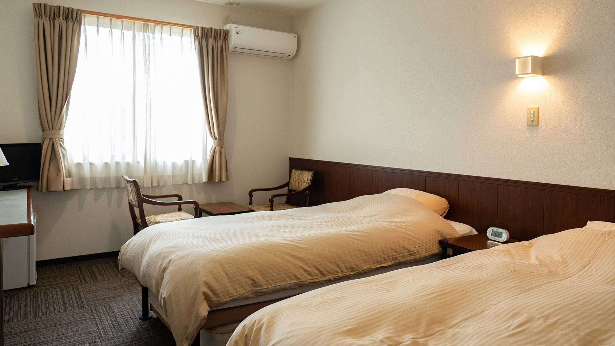 Une chambre d’hôtel avec deux lits, un bureau, une chaise, un appareil de climatisation et un luminaire fixé au mur.