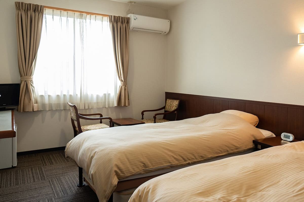 A hotel room with two beds, a desk, a chair, an air conditioning unit, and a wall-mounted light.