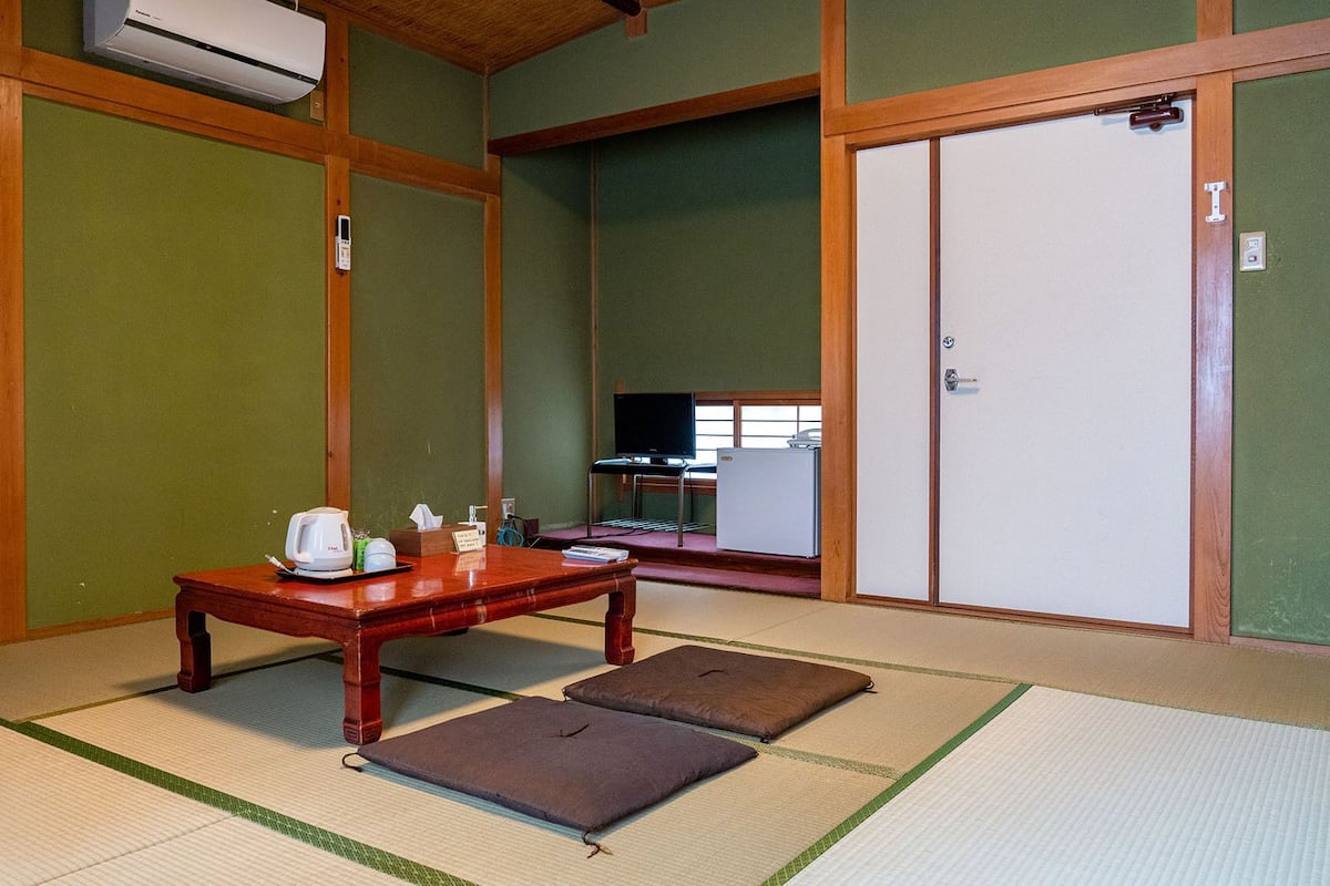 A traditional Japanese room with tatami flooring, a wooden table with a kettle, a television, and a refrigerator.