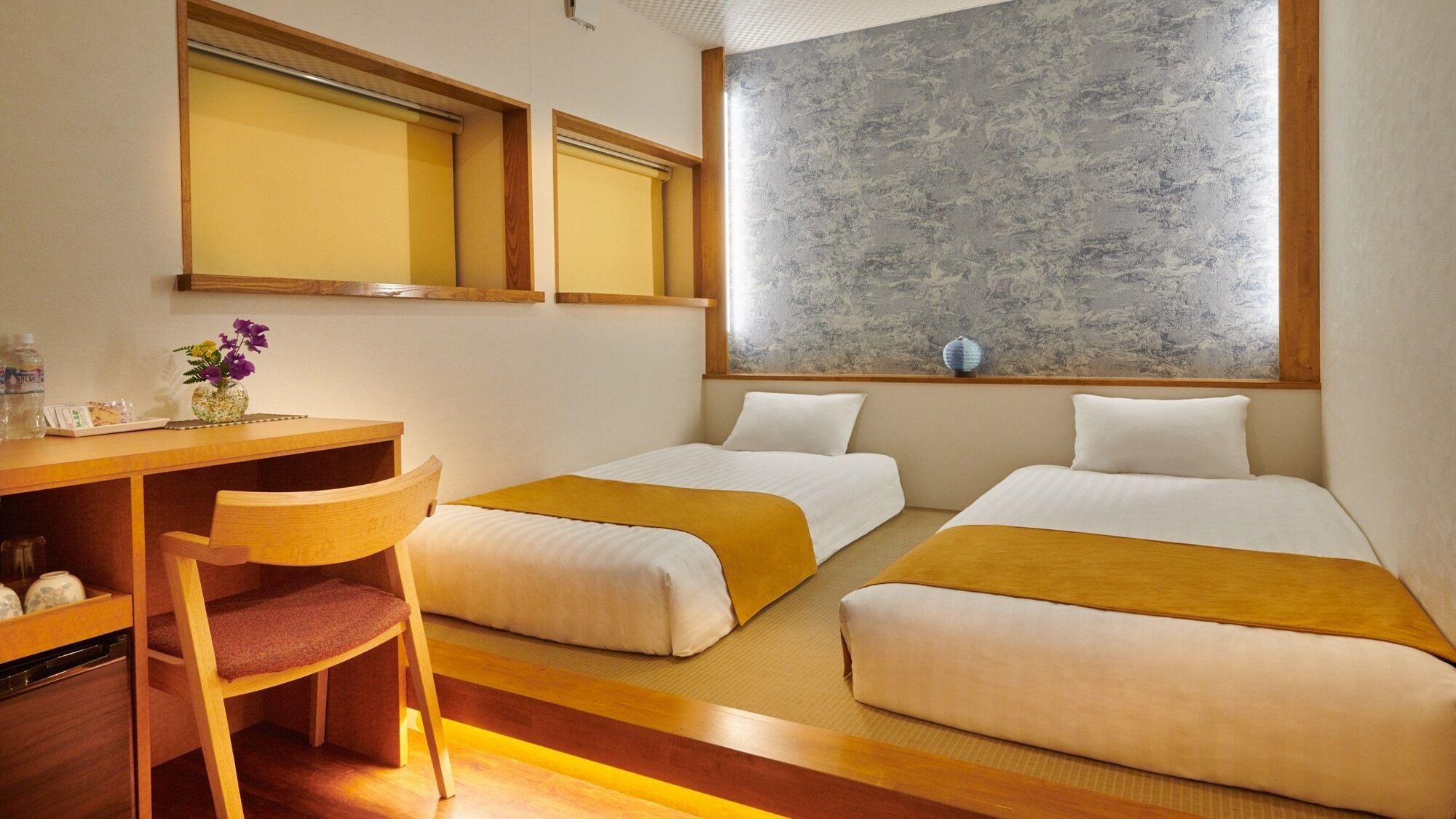 A hotel room with two beds, a desk, a chair, a small table with a vase of flowers, and a large window with a textured wall.