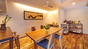 Uma sala de jantar com mesa de madeira, cadeiras, um vaso com flores, um quadro na parede e uma estante com utensílios de cozinha.