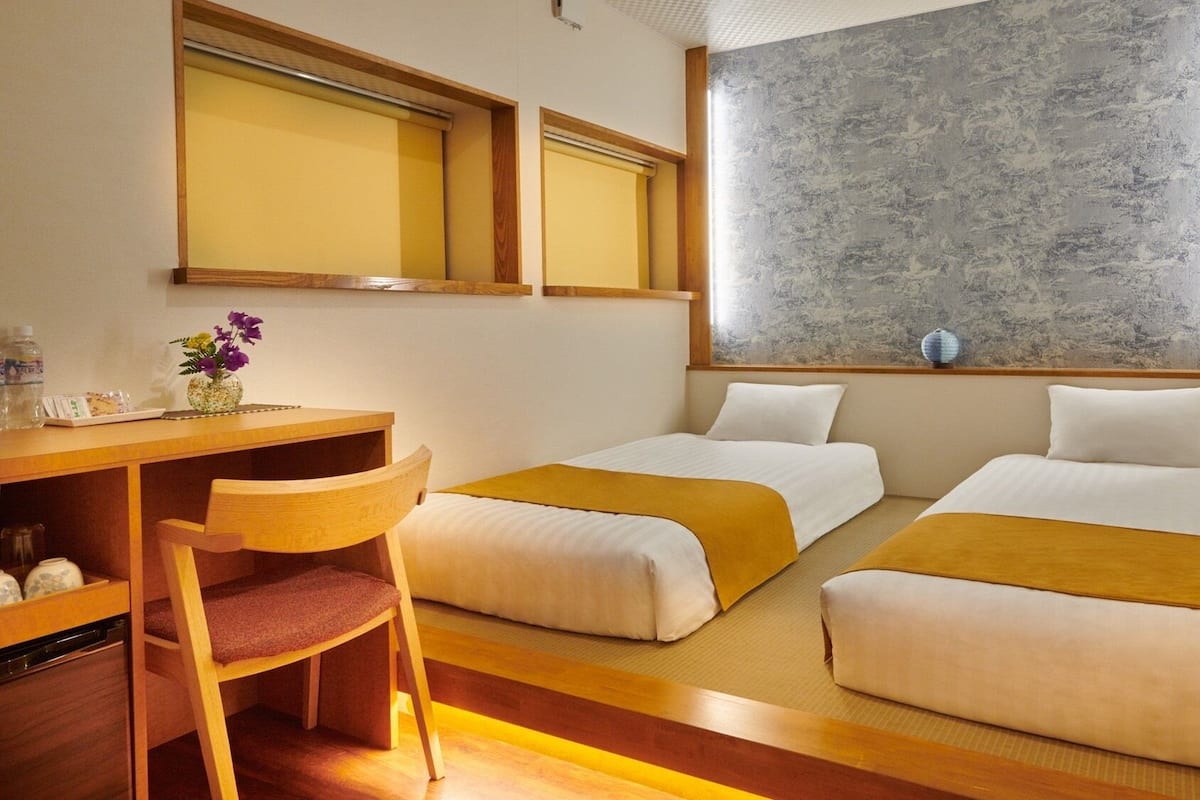 Quarto de hotel com duas camas, uma escrivaninha, uma cadeira, uma mesa pequena com um vaso de flores e uma janela grande com parede texturizada.