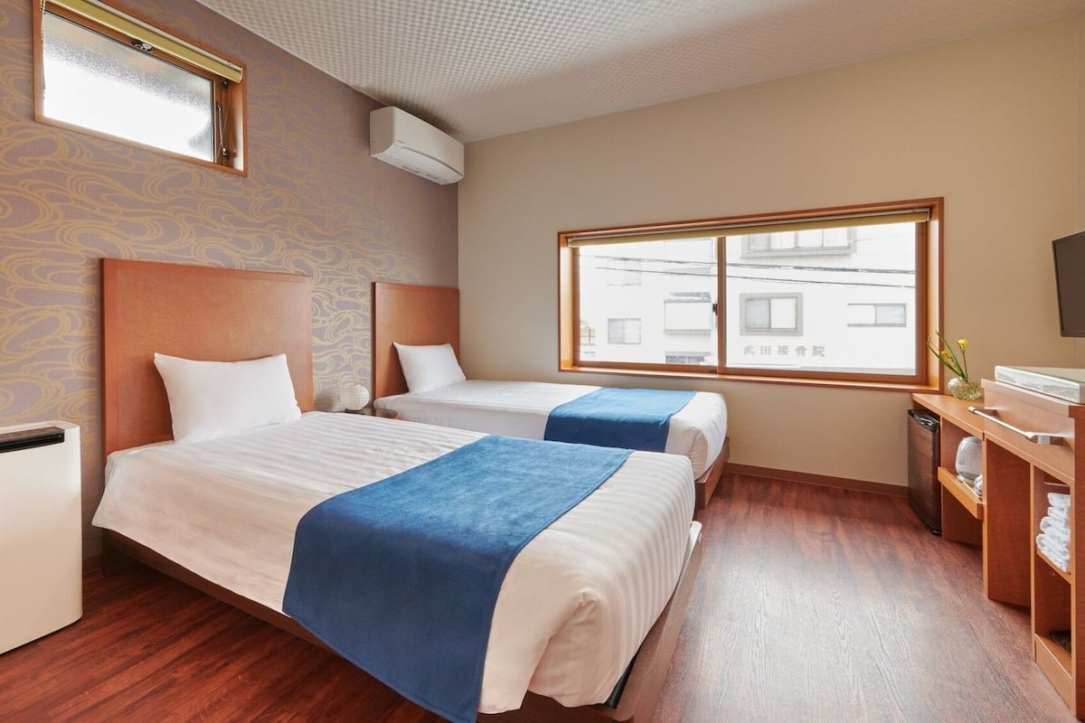 Quarto de hotel com duas camas, uma cabeceira de madeira, uma janela com vista, mesa de cabeceira e televisão.