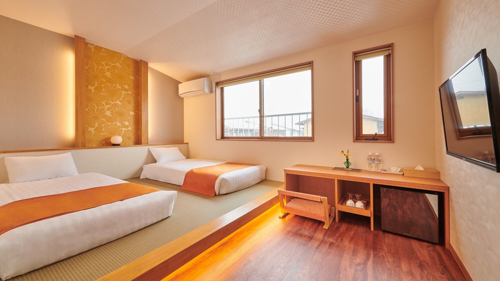 A hotel room with two beds, a wooden desk, a TV, and large windows.