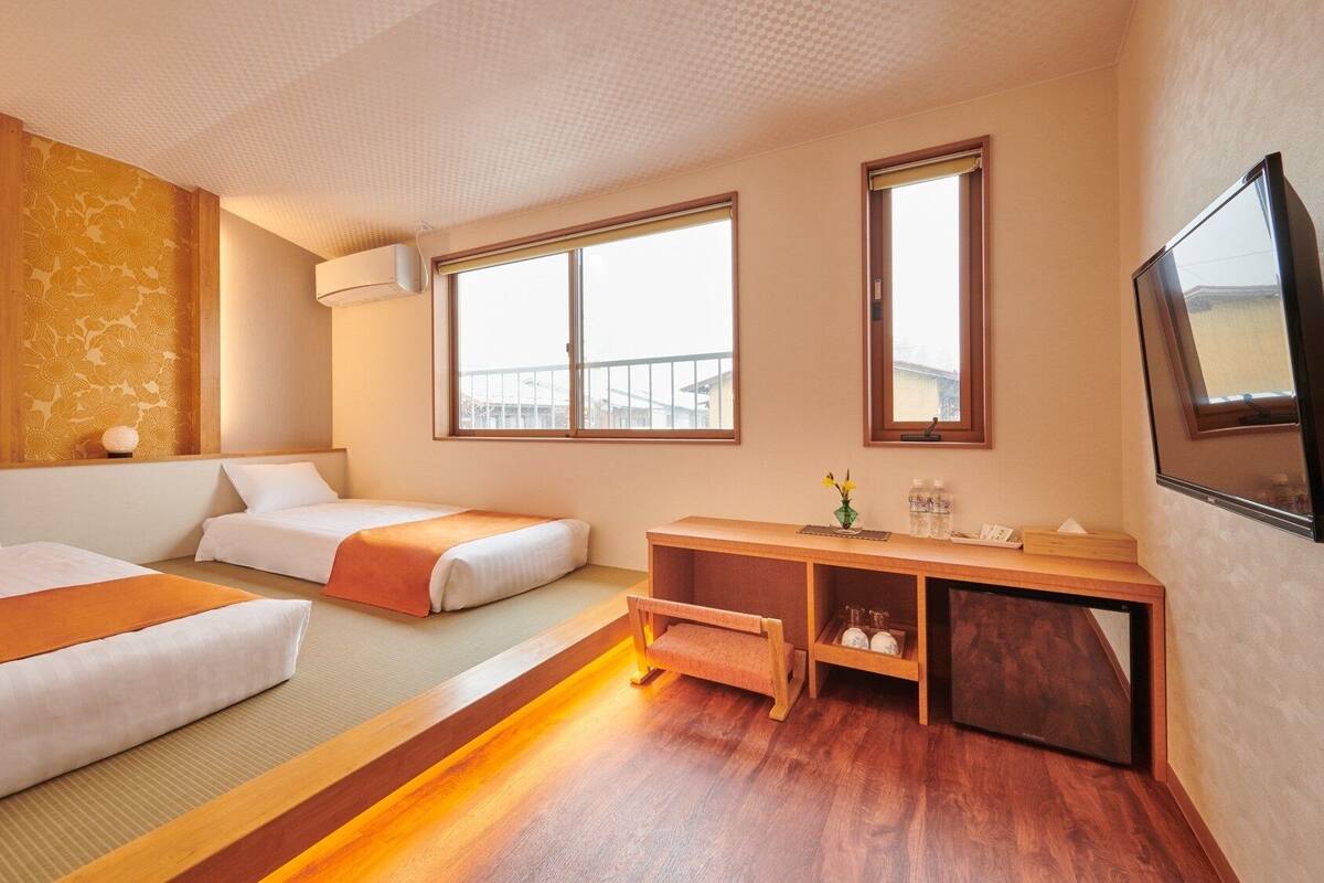 Quarto de hotel com duas camas, uma secretária de madeira, televisão e janelas amplas.