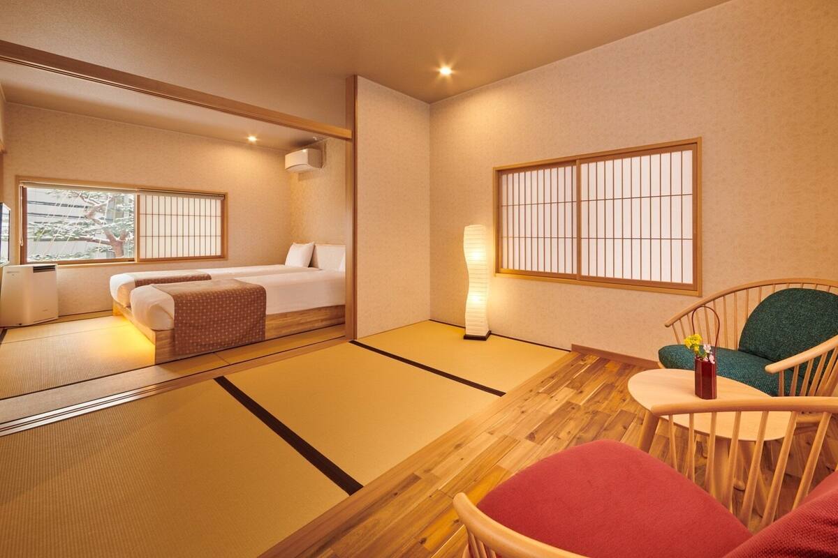 Quarto de hotel com piso de tatame, cama futon, cadeira de madeira, pequena mesa com um vaso e janela com tela shoji.