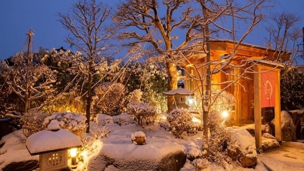 Um edifício japonês tradicional com estrutura de madeira e um emblema vermelho, cercado por árvores e pedras cobertas de neve.