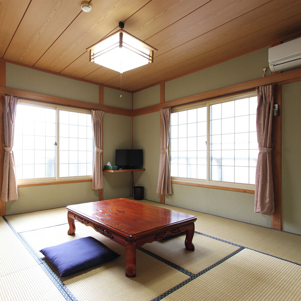 A traditional Japanese room with tatami flooring, a wooden table, a television on a shelf, and large windows with curtains.