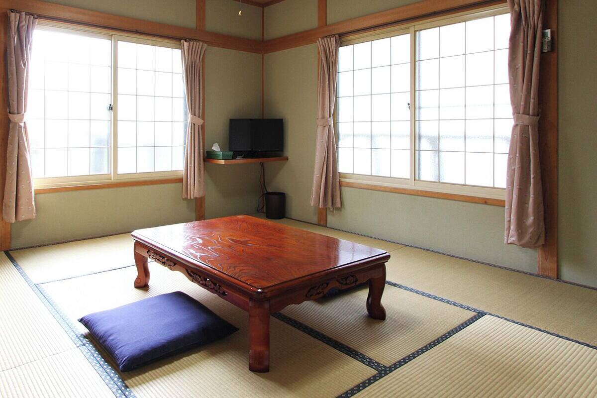 A traditional Japanese room with tatami flooring, a wooden table, a television on a shelf, and large windows with curtains.