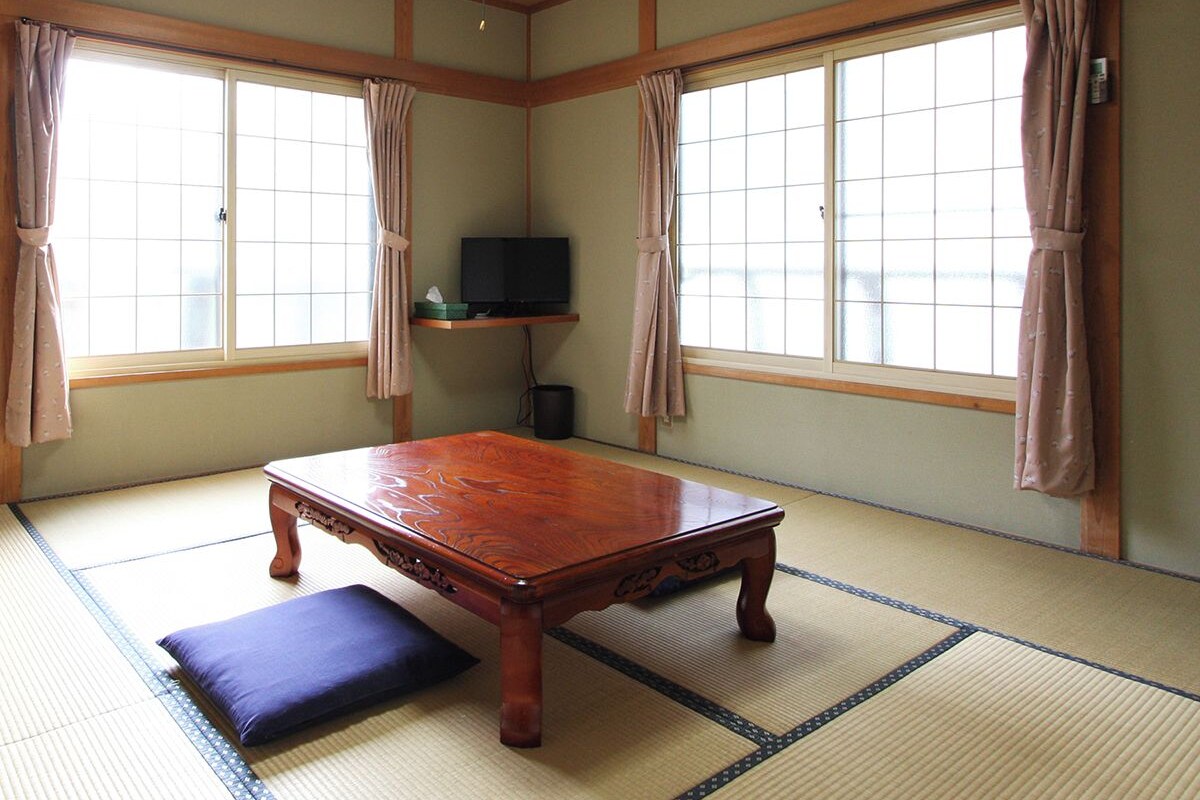 A traditional Japanese room with tatami flooring, a wooden table, a television on a shelf, and large windows with curtains.