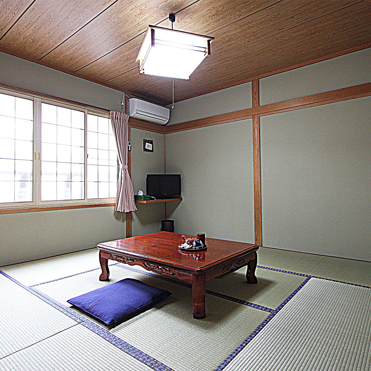 A traditional Japanese room with tatami flooring, a low wooden table, a television on a shelf, and a ceiling fan.