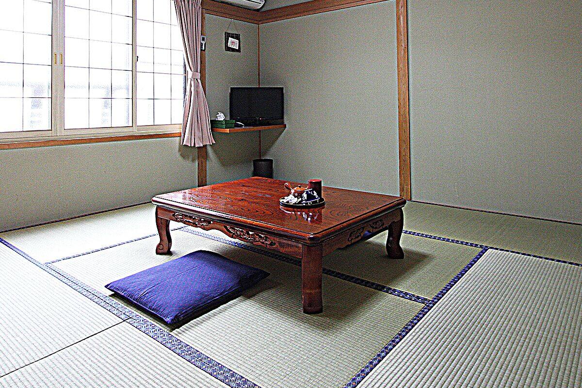 A traditional Japanese room with tatami flooring, a low wooden table, a television on a shelf, and a ceiling fan.