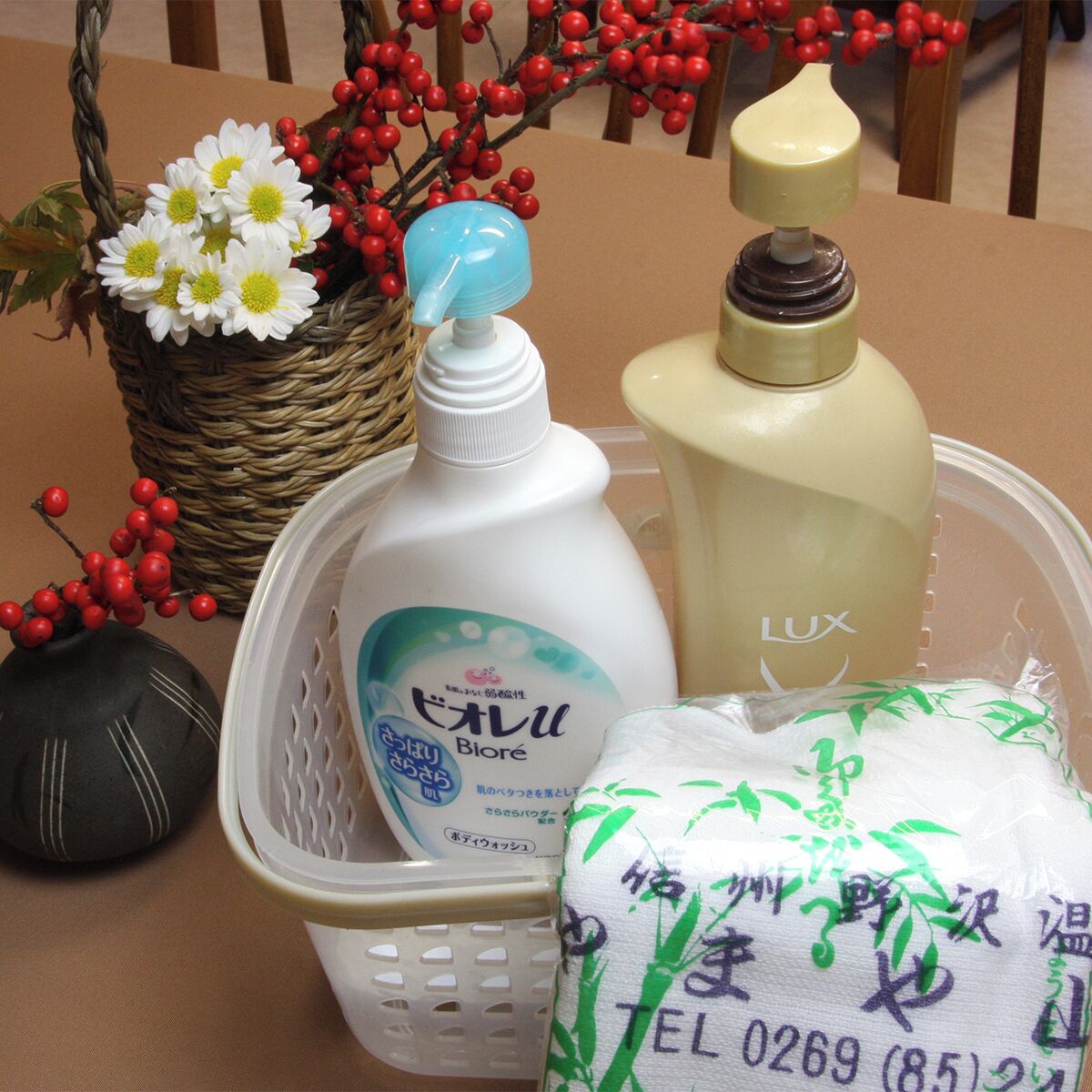 A table with a basket containing a bottle of Biore soap, a bottle of Lux lotion, a package of tissue, and a decorative vase with flowers and berries.