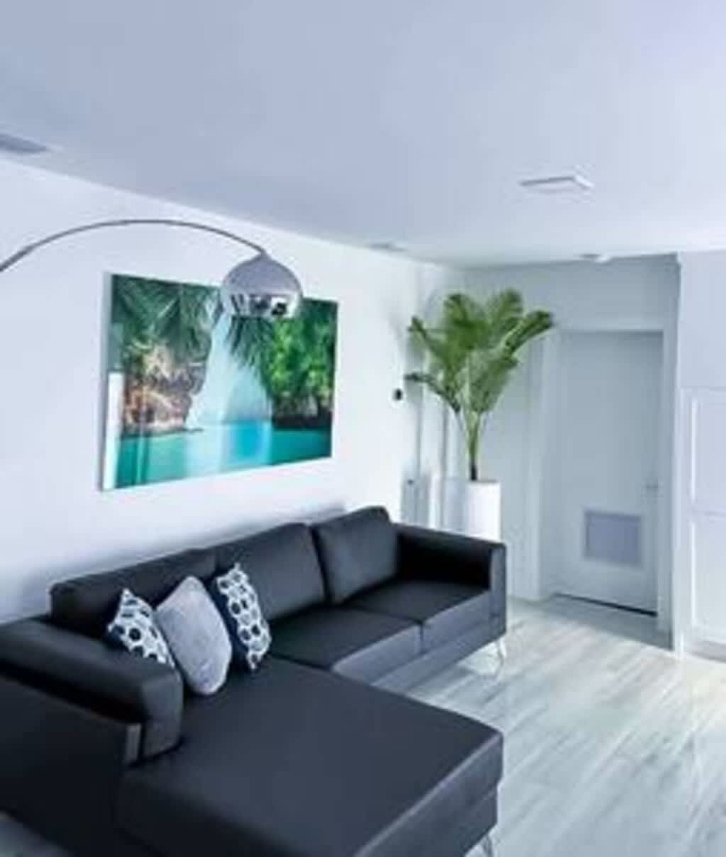 A modern living room with a black sofa, a wall-mounted lamp, a large framed picture of a tropical scene, and a potted plant.