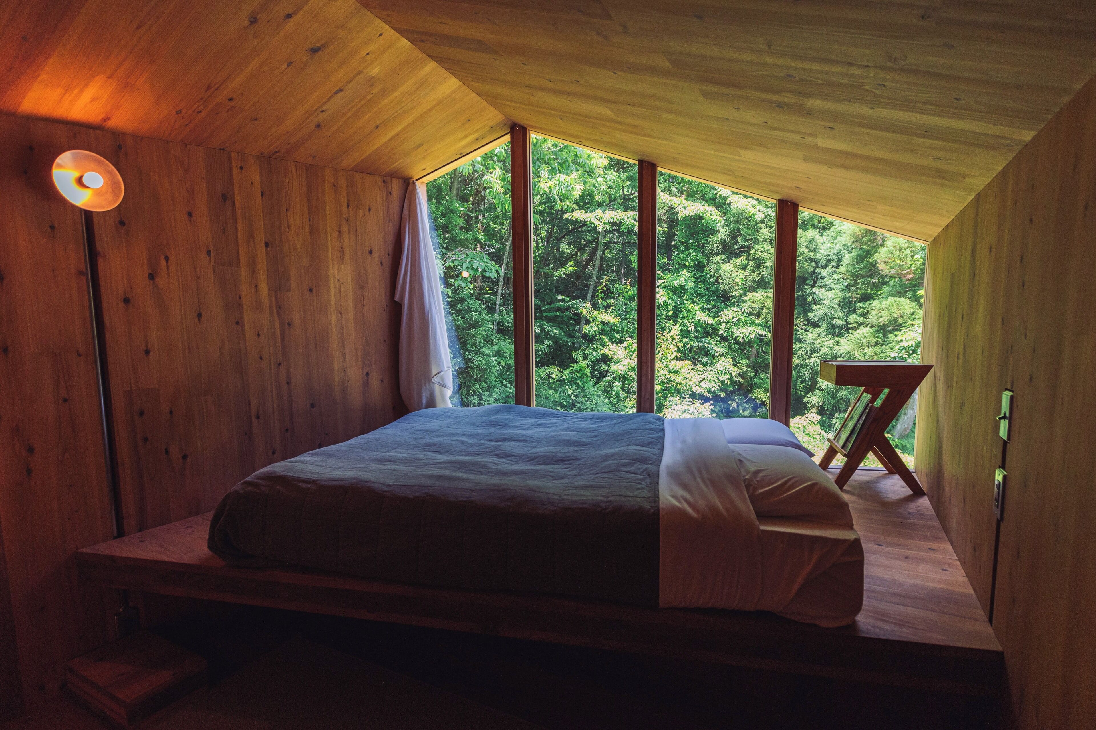 Een slaapkamer met een houten plafond en grote ramen, een bed, een nachtkastje en een vloerlamp.