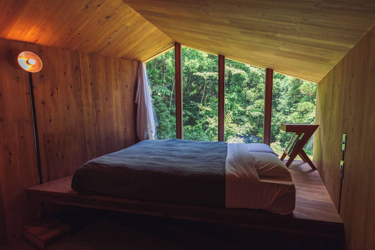 Un dormitorio con techo de madera y ventanas amplias, una cama, una mesita de noche y una lámpara de pie.