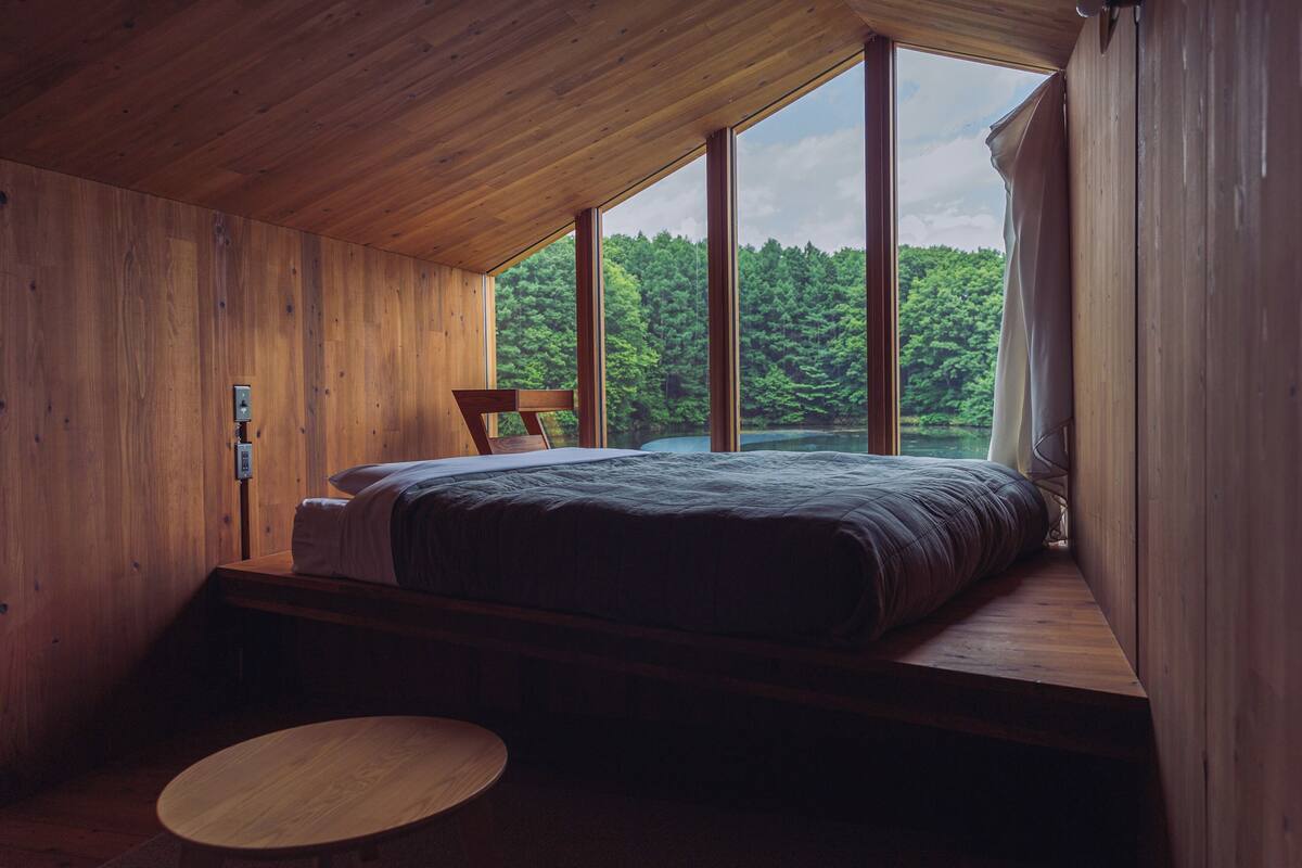 Un dormitorio con una cama grande, una mesa de madera y vistas a un bosque a través de amplios ventanales.