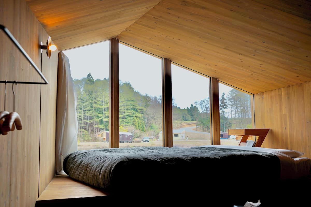 Un dormitorio con un ventanal grande, paredes de madera y una cama con colcha oscura.