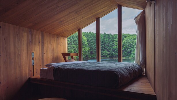 Ein Schlafzimmer mit einem großen Bett, einem Holztisch und Blick auf einen Wald durch große Fenster.