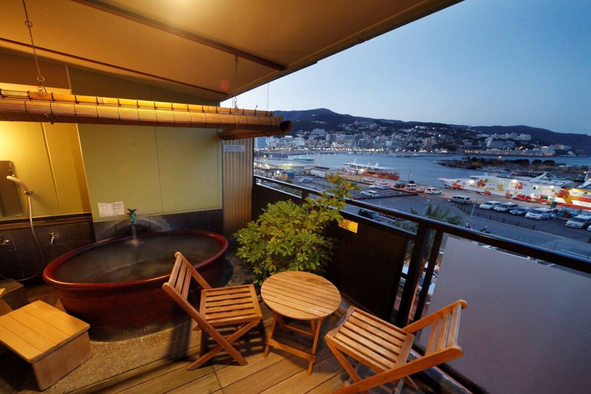 Un balcon doté d’un jacuzzi, de chaises en bois, d’une petite table et offrant une vue sur un port avec des bateaux et des bâtiments.