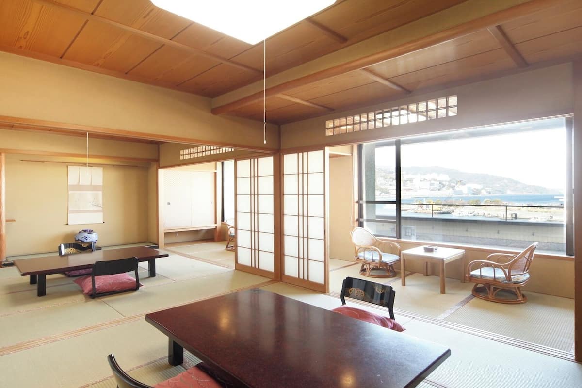 Une pièce de style japonais traditionnel, avec un sol recouvert de tatamis, une table basse, des chaises et une vue sur un paysage côtier.
