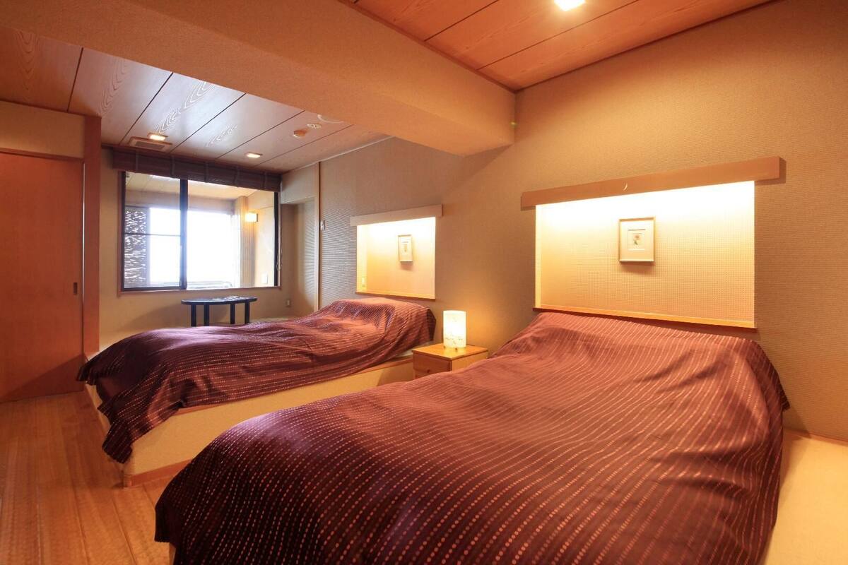 Une chambre d’hôtel avec deux lits, une petite table, un miroir et une fenêtre laissant entrer la lumière naturelle.