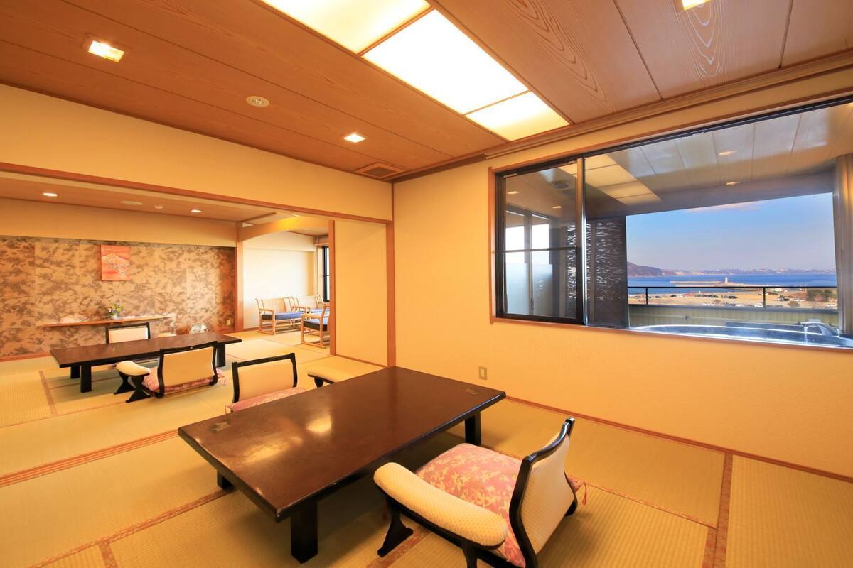 Une pièce de style japonais traditionnel, avec un sol recouvert de tatamis, une table basse et des chaises, une grande fenêtre donnant sur la plage et une porte coulissante menant à une autre pièce.