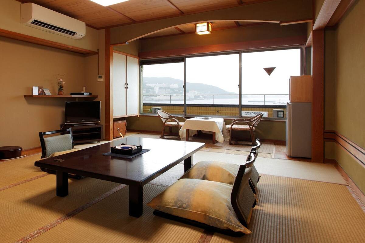 Une pièce de style japonais traditionnel, avec un sol recouvert de tatamis, une table basse, des chaises, un téléviseur et une vue sur la ville grâce à de grandes fenêtres.