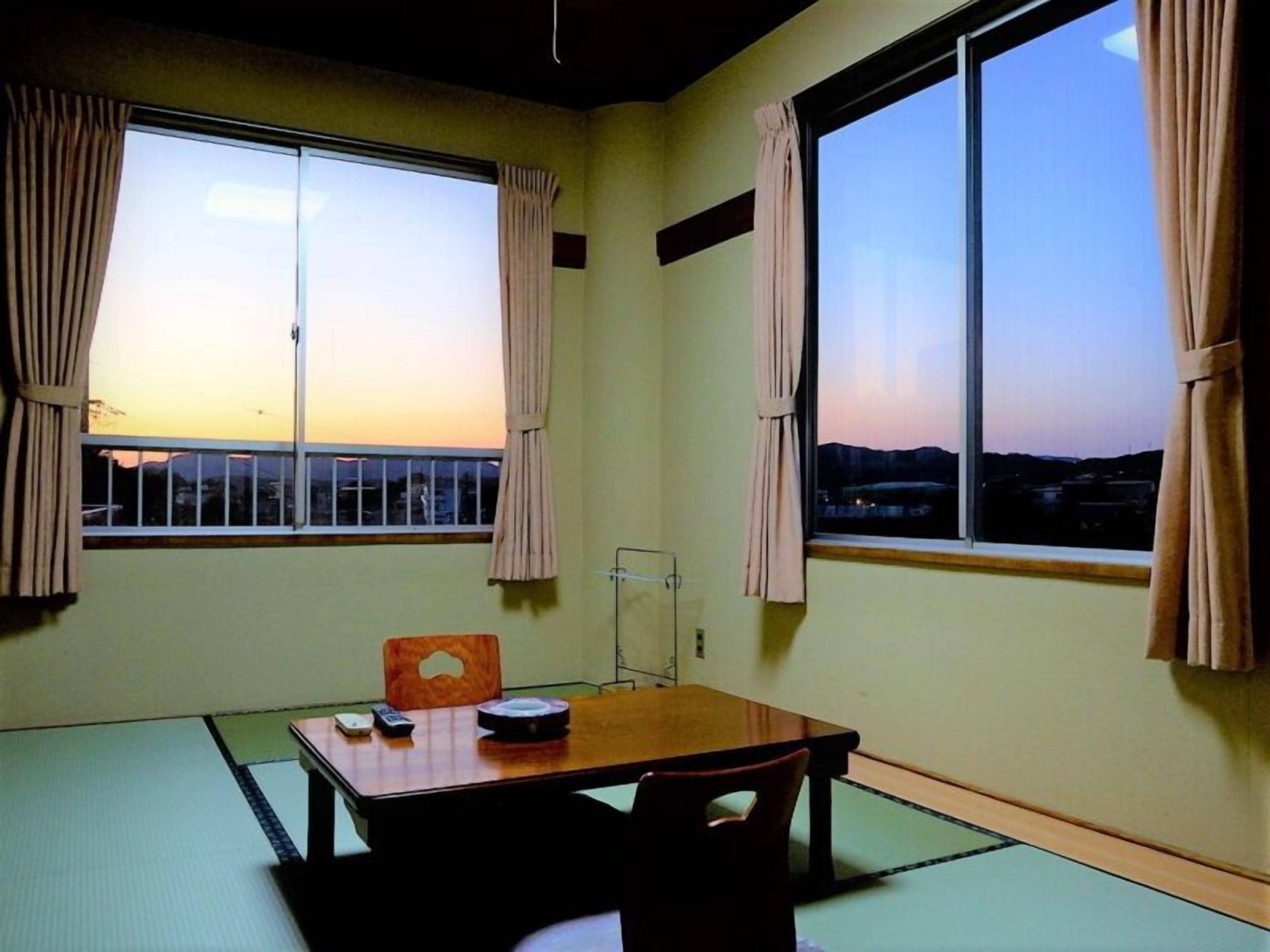 Quarto com piso de tatame, uma mesa de madeira com um controle remoto e vista para o pôr do sol através de grandes janelas.